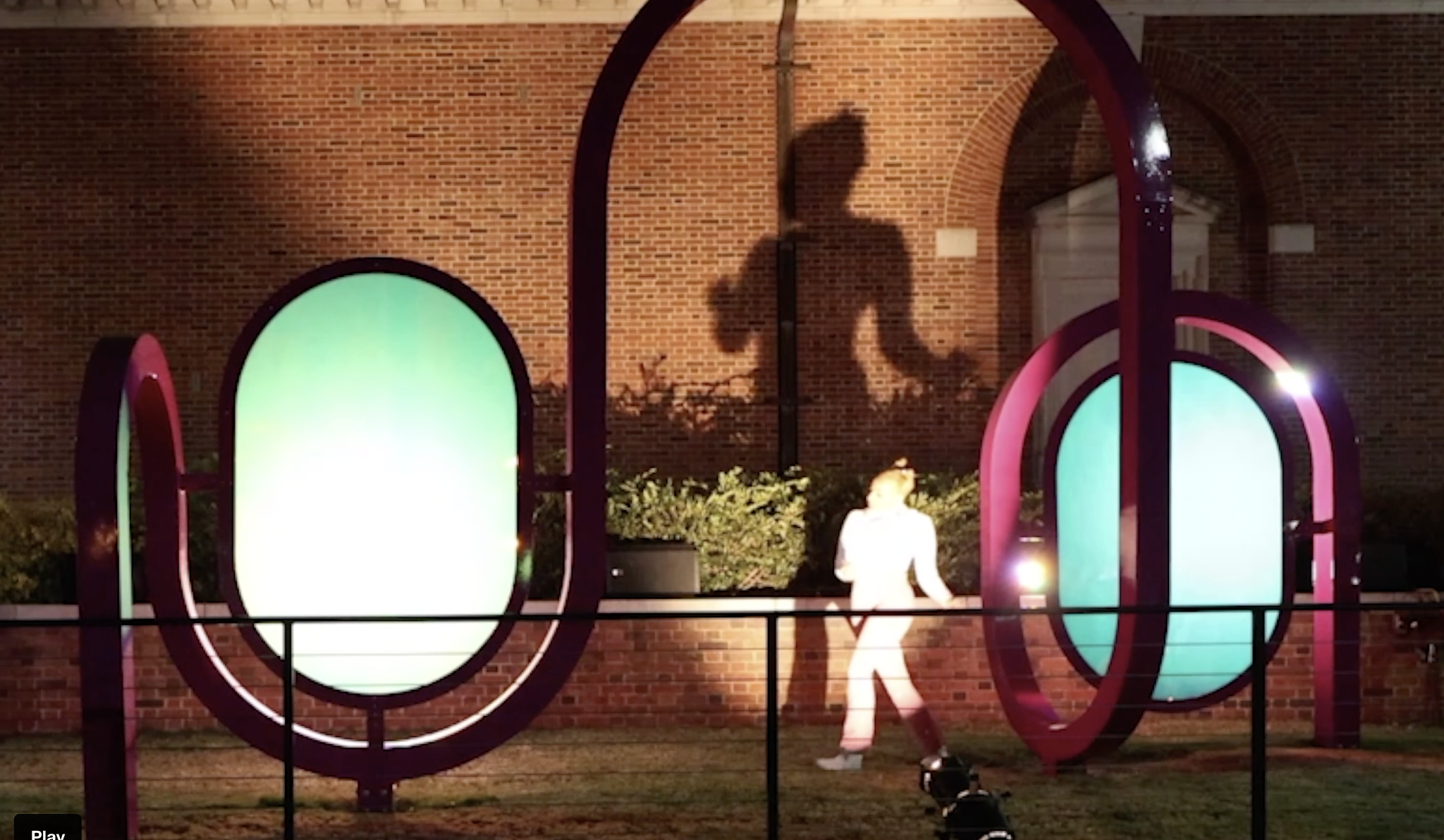 A dancer performs in front of a light outside at night, casting her shadow on a brick wall